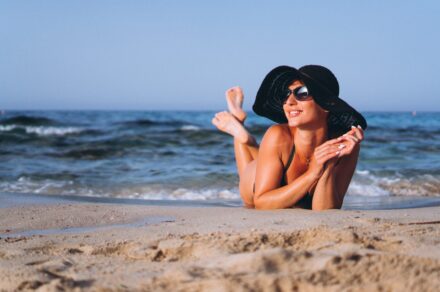 Femme avec un chapeau et des lunettes de soleil en train de bronzer sur le sable au bord de l'eau